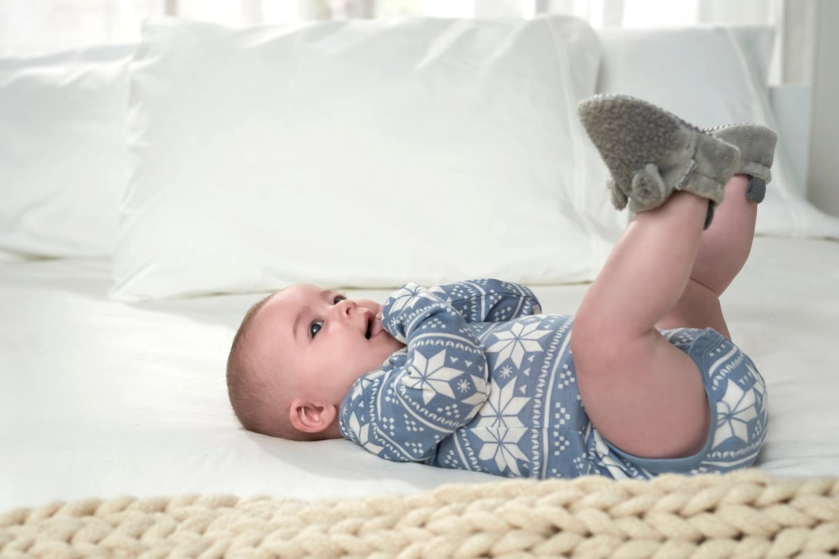 Baby lying on a white blanket wearing a blue patterned outfit and gray mittens.