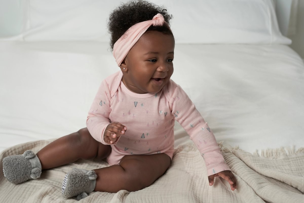 Baby in a pink outfit with a headband sitting on a white blanket