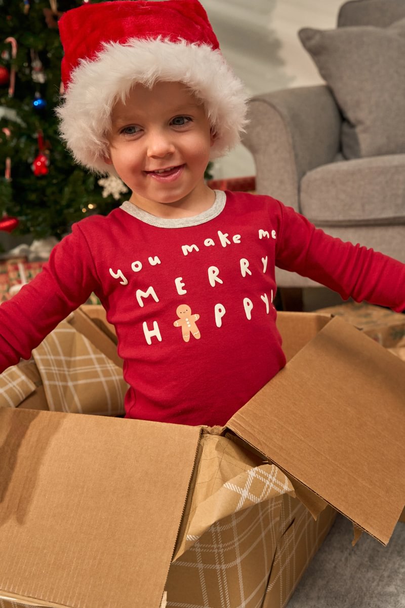 Child wearing a red Christmas shirt and Santa hat, standing in front of a decorated Christmas tree.