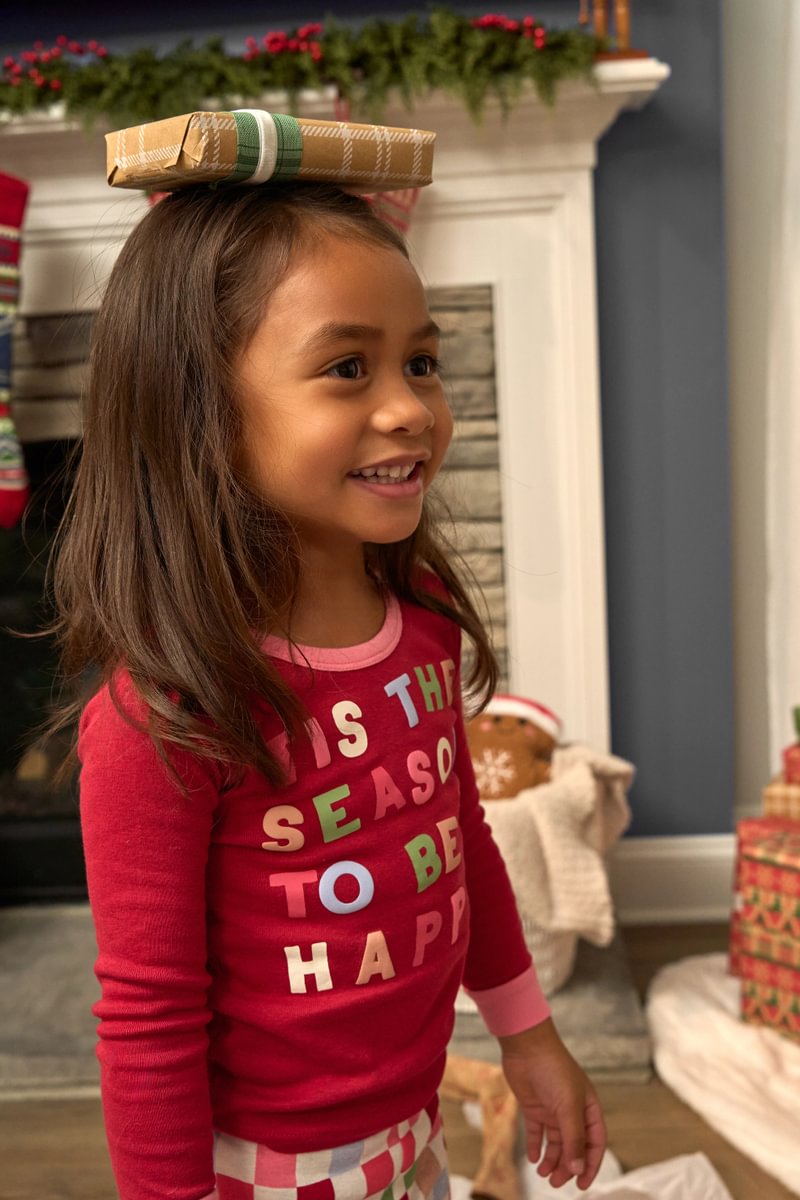 Toddler girl wearing a red shirt with Christmas-themed text, standing in a festive room with a fireplace and wrapped gifts.