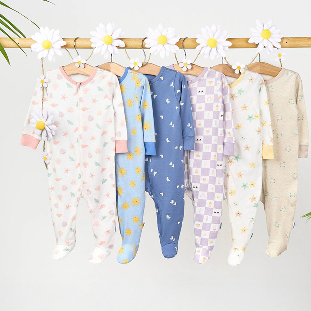 Colorful baby and toddler footed sleepwear from Gerber Childrenswear’s $5 Frenzy Sale, featuring playful summer-themed prints like suns, seashells, butterflies, and rainbows, all hanging from a daisy-decorated bamboo rod with palm leaves, showcasing soft and cozy one-piece pajamas perfect for warm-weather nights.