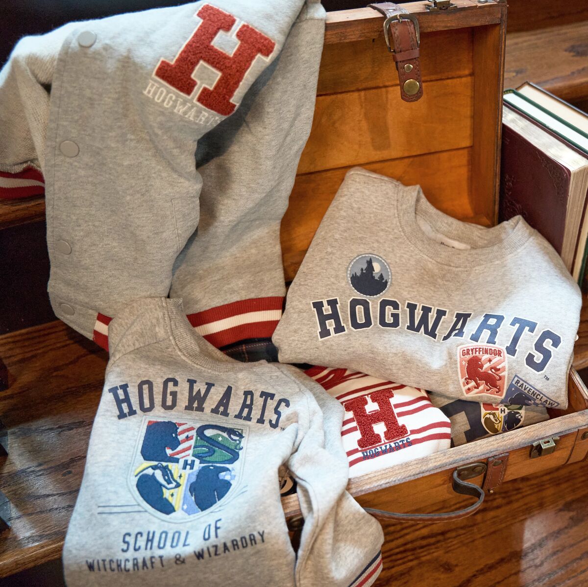 A collection of gray and red Hogwarts-themed apparel, including a varsity jacket and sweatshirts, arranged inside and around a wooden trunk.