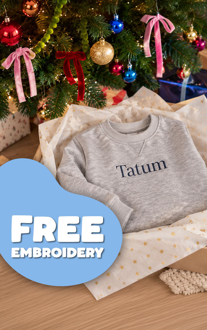 A light gray crewneck sweatshirt with the name 'Tatum' embroidered in navy on the chest, displayed in a gift box with tissue paper beneath a decorated Christmas tree. A blue graphic overlay in the bottom left reads 'FREE EMBROIDERY'.