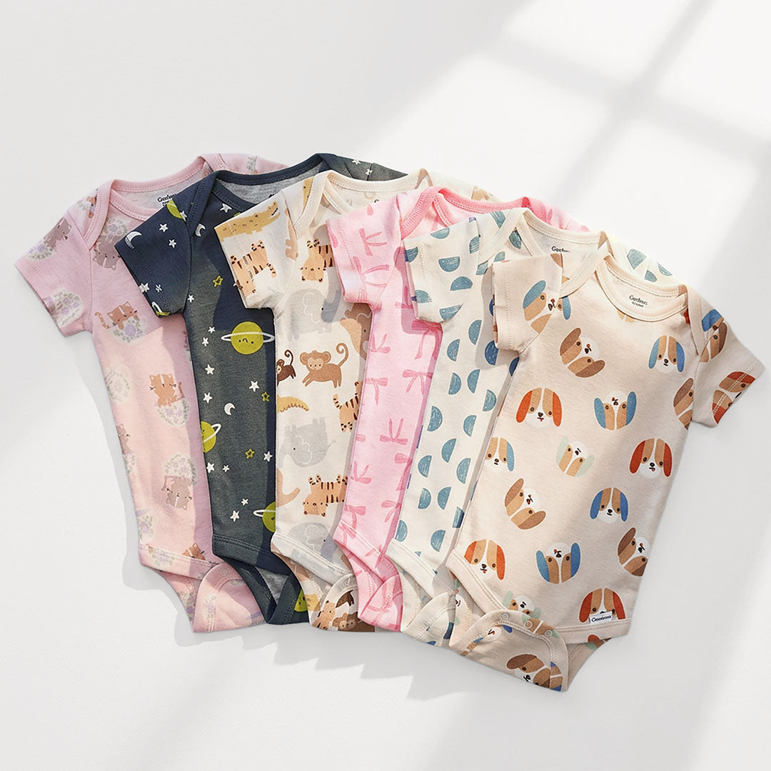 A stack of sixe baby bodysuits in various pastel colors, each featuring different cute cartoon prints like planets, animals, and musical notes, set against a clean white background.