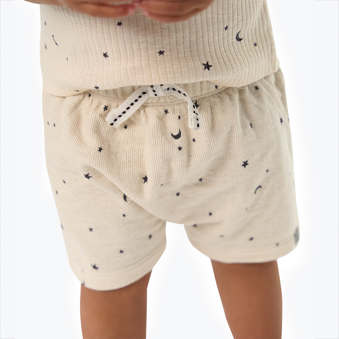 Close-up of a child wearing cream-colored ribbed knit shorts and matching top with small black star and crescent moon prints, against a plain white background.