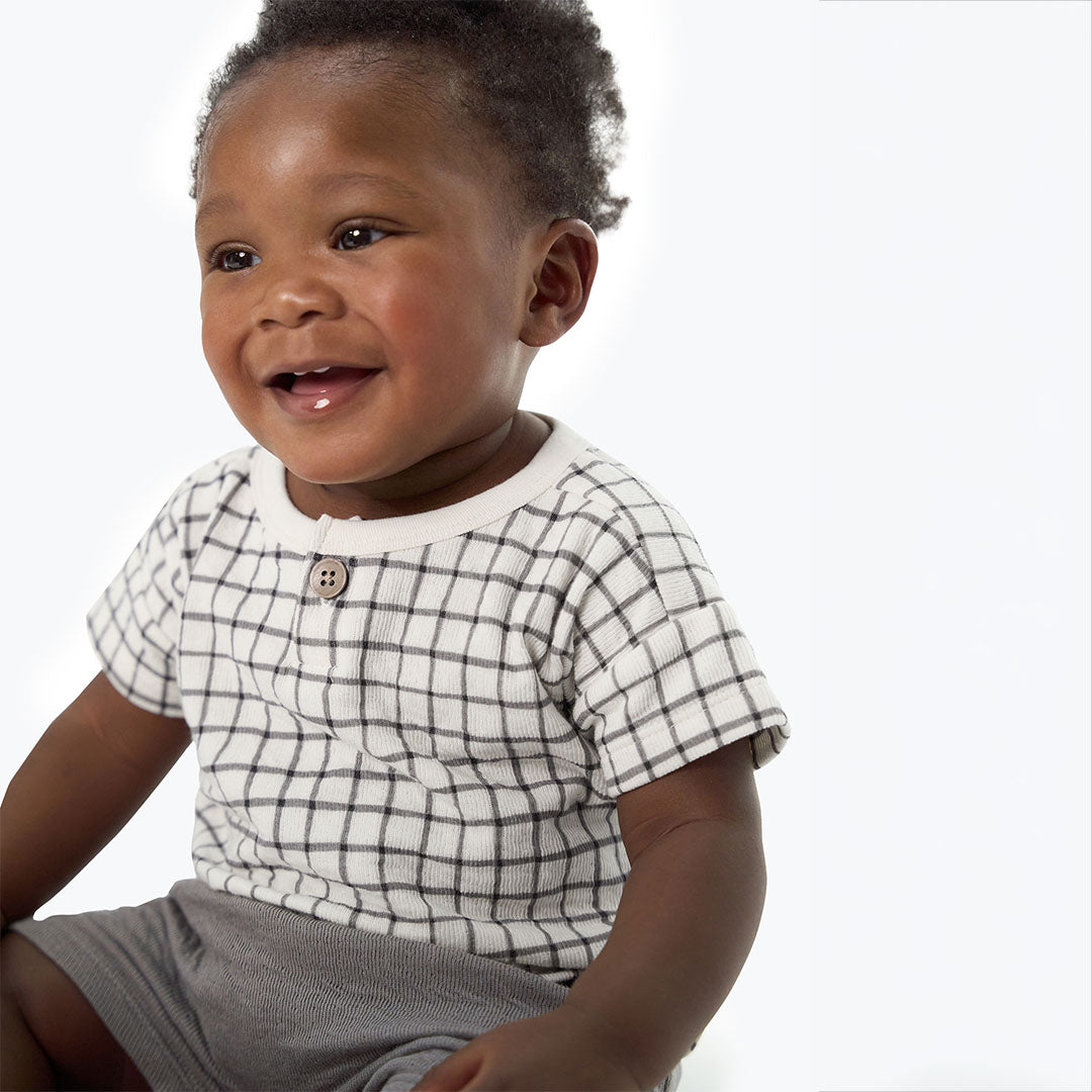 A smiling baby wearing a white short-sleeve henley shirt with a black grid pattern and gray shorts, photographed against a clean white background.