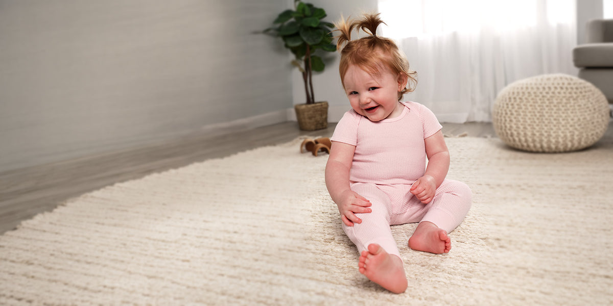 A smiling baby girl with her hair in two buns sits on a cream rug wearing a light pink short-sleeve bodysuit and pants set. The room has light gray walls, a white curtain, a potted plant, and a round knitted pouf.