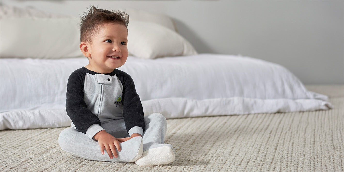 A toddler sits cross-legged on a textured cream rug in a bedroom, wearing a gray and black long-sleeve zippered pajamas.