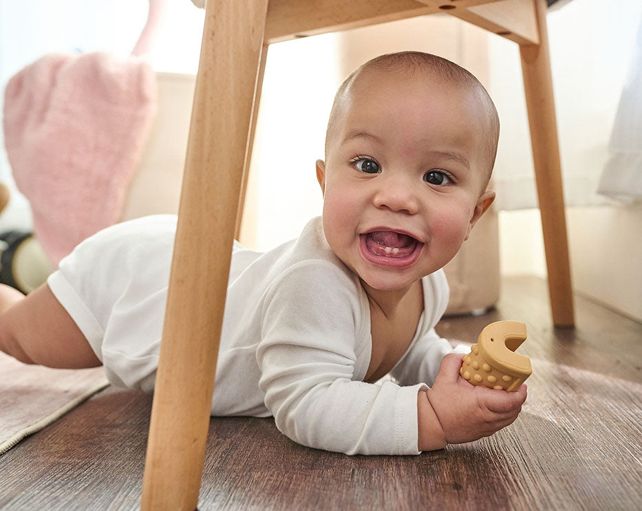 A smiling baby lying on a wooden floor under a wooden table, wearing a white long-sleeve bodysuit and holding a small beige teething toy.