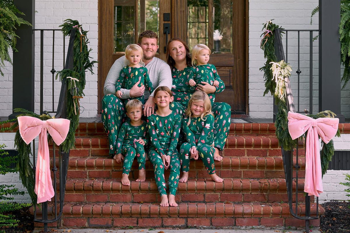 A family of six wearing matching green Christmas-themed pajamas with a red and white pattern, posed together on the brick steps of a house decorated with green garlands and pink bows.