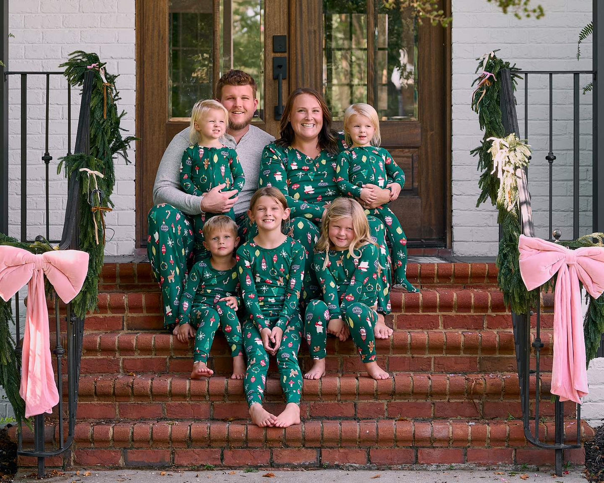 A family of seven wearing matching green holiday-themed pajamas with a festive print, posing together on the brick steps of a decorated house porch.