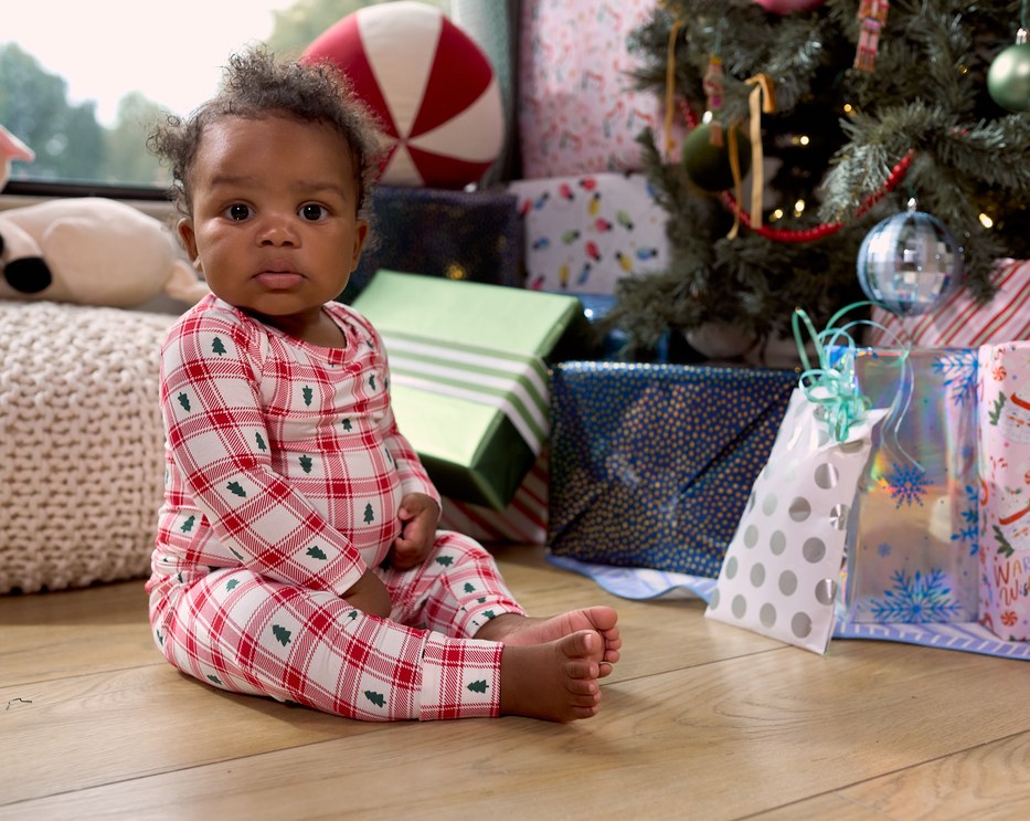 A baby sitting on a wooden floor in front of a Christmas tree and wrapped gifts, wearing a red and white plaid outfit set with small green tree prints.