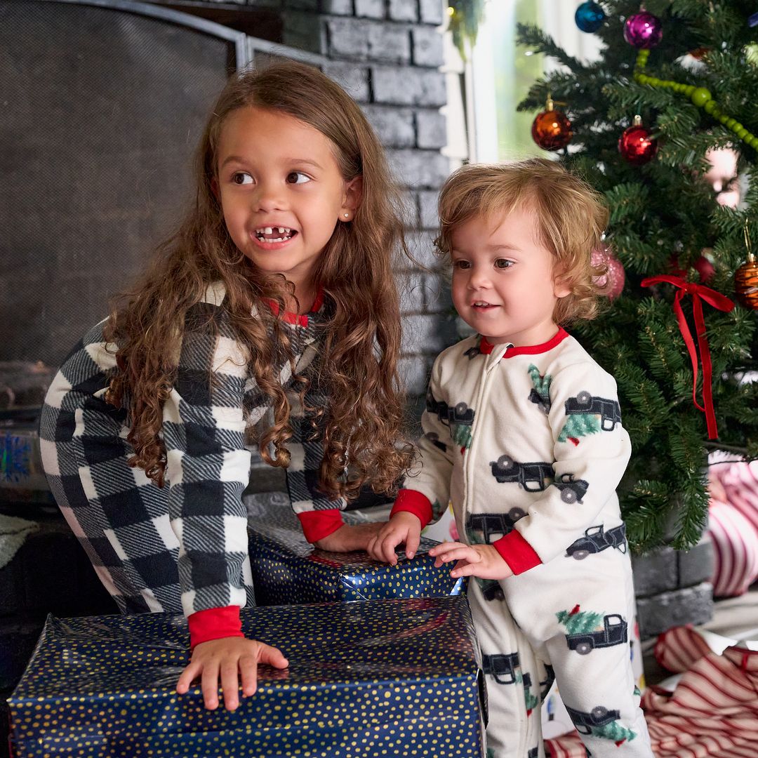 Two children in holiday pajamas sit beside a decorated Christmas tree and wrapped gifts.