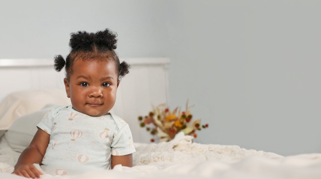 Baby in a light blue onesie sitting on a bed with an adult next to them, with a neutral background.