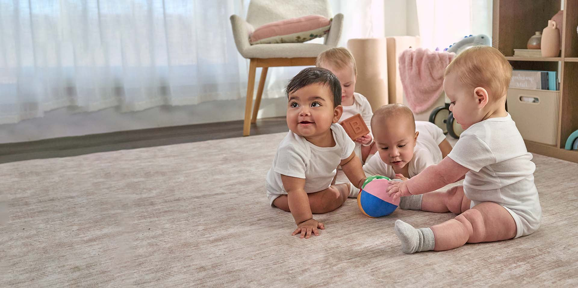 Four babies playing with toys on a wooden floor in a home setting.