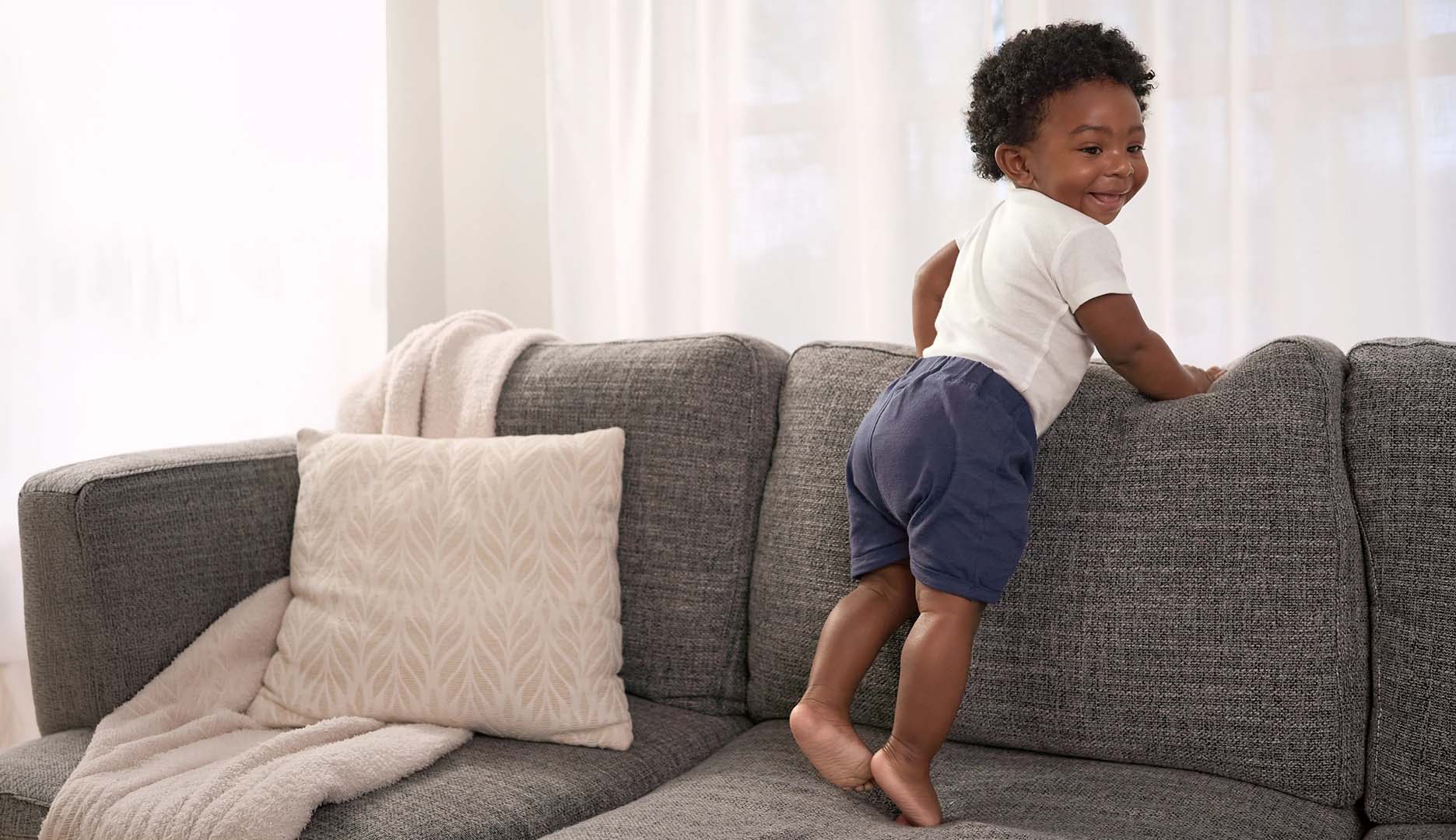Child standing on a gray sofa with white curtains in the background