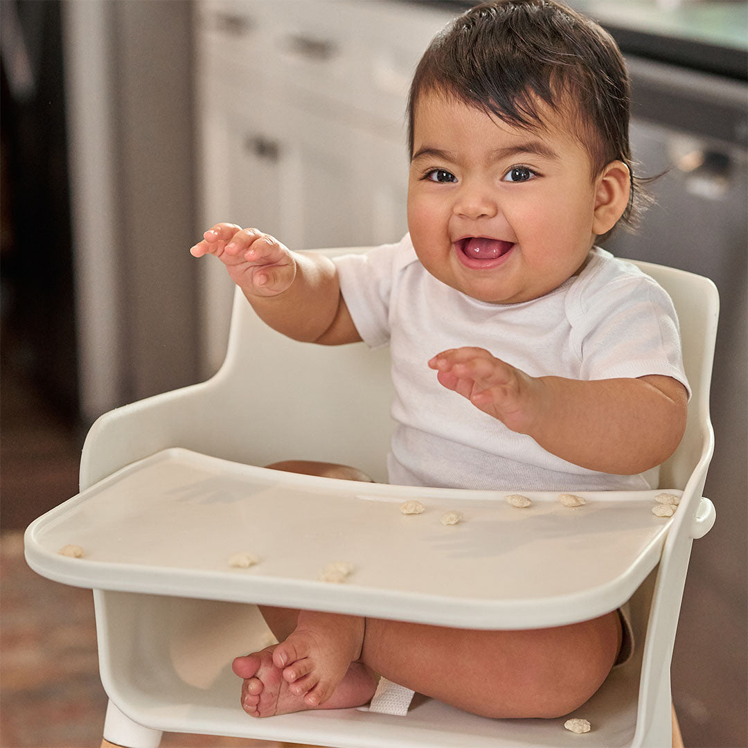 A smiling baby sitting in a white high chair with a tray, wearing a white baby bodysuit in a kitchen setting.