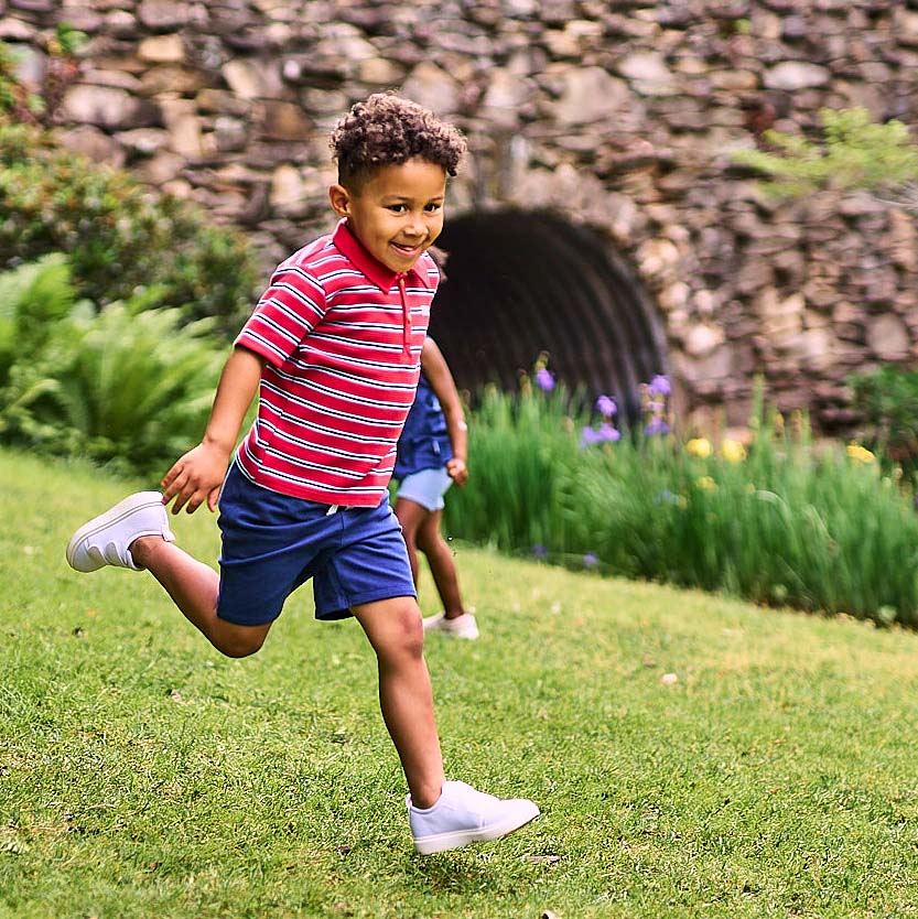 A young boy running across a grassy lawn wearing a red and white striped polo shirt, blue shorts, and white sneakers, with another child blurred in the background near a stone wall and greenery.