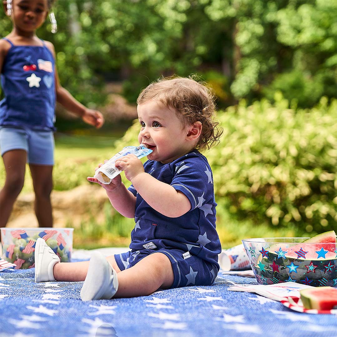 A toddler wearing a navy blue short-sleeve romper with white stars sits on a blue picnic blanket outdoors, holding a food pouch. Another child in a matching navy top is blurred in the background on a sunny day.