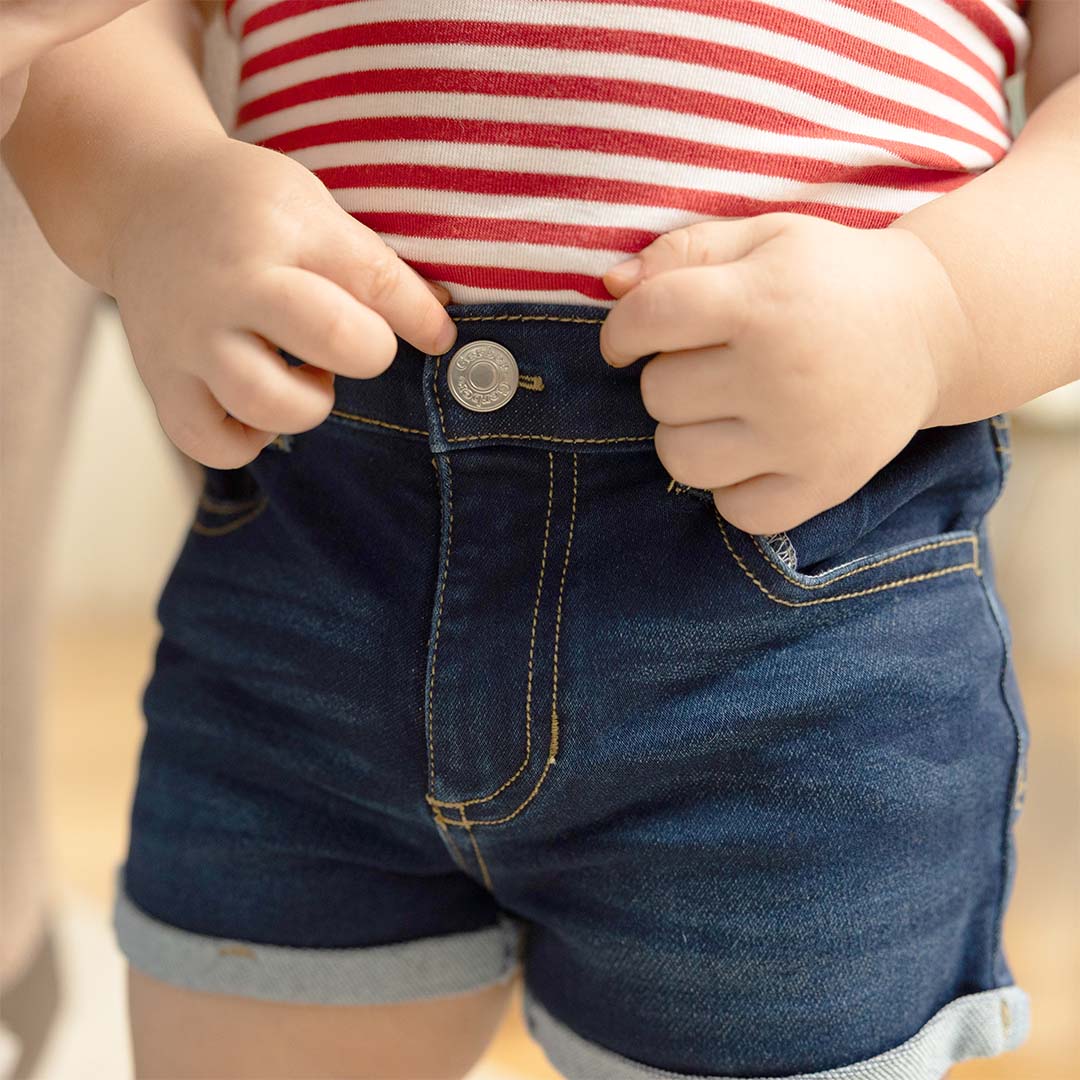 Close-up of a child wearing dark blue denim shorts with rolled hems and a red-and-white striped top, holding the shorts closed with both hands.