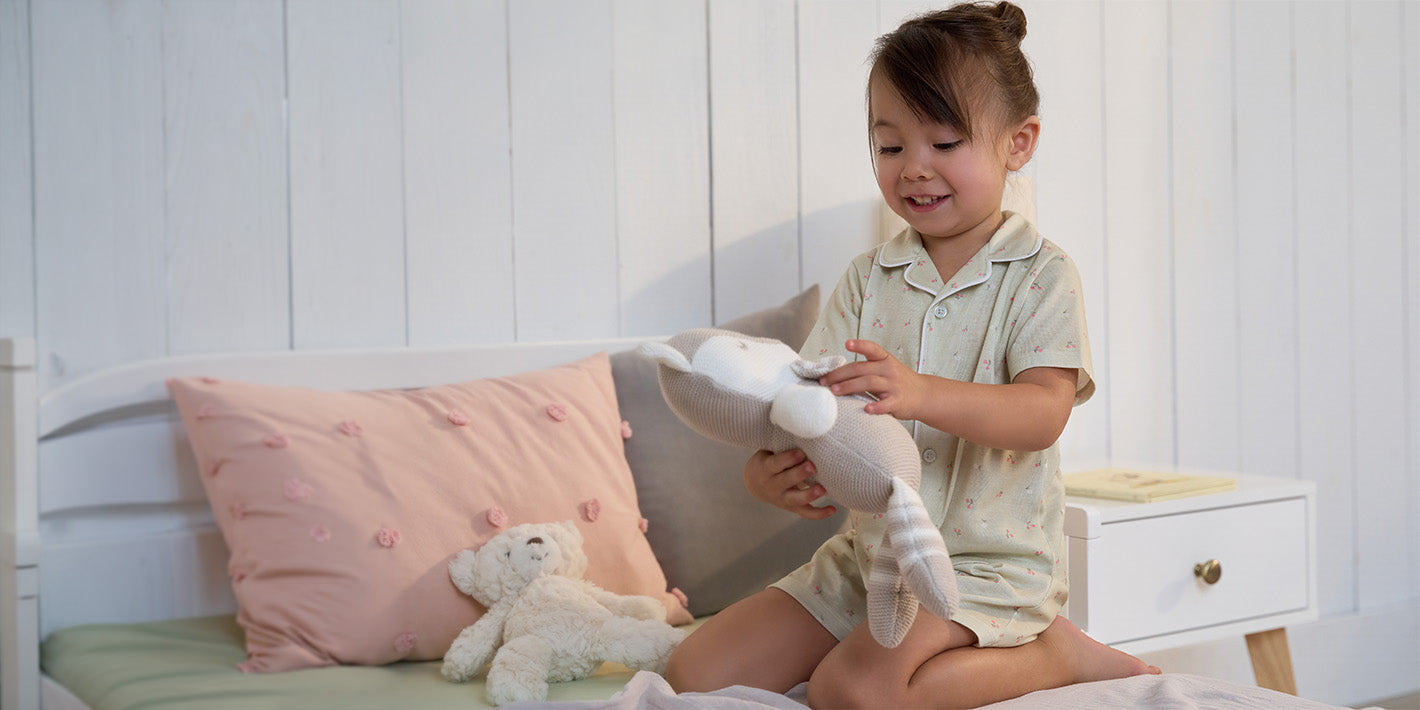 A young child wearing light green pajamas sits on a bed holding a white plush stuffed animal, with a pink pillow and a small teddy bear on the bed beside them.