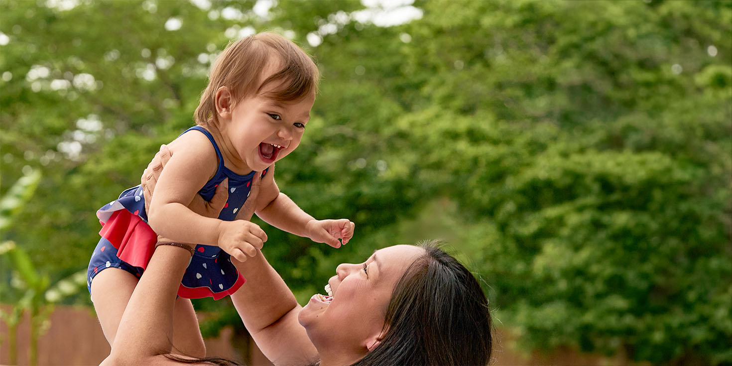A baby wearing a patriotic red, white, and blue swimsuit is being held up by an adult woman's arms in an outdoor backyard setting with green trees in the background.