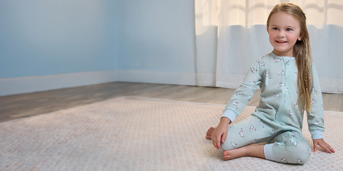 Young girl in light blue pajamas sitting on a rug in a softly lit room.