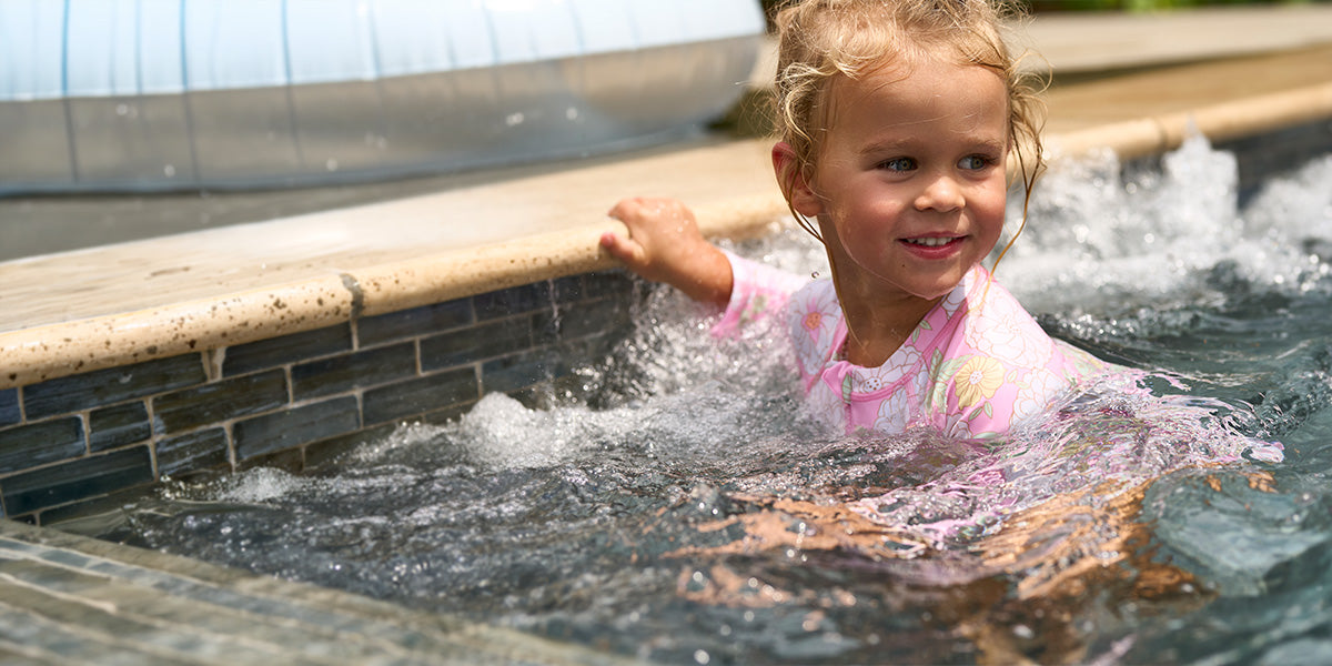 Child in a pink swimsuit standing by a pool with a garden background