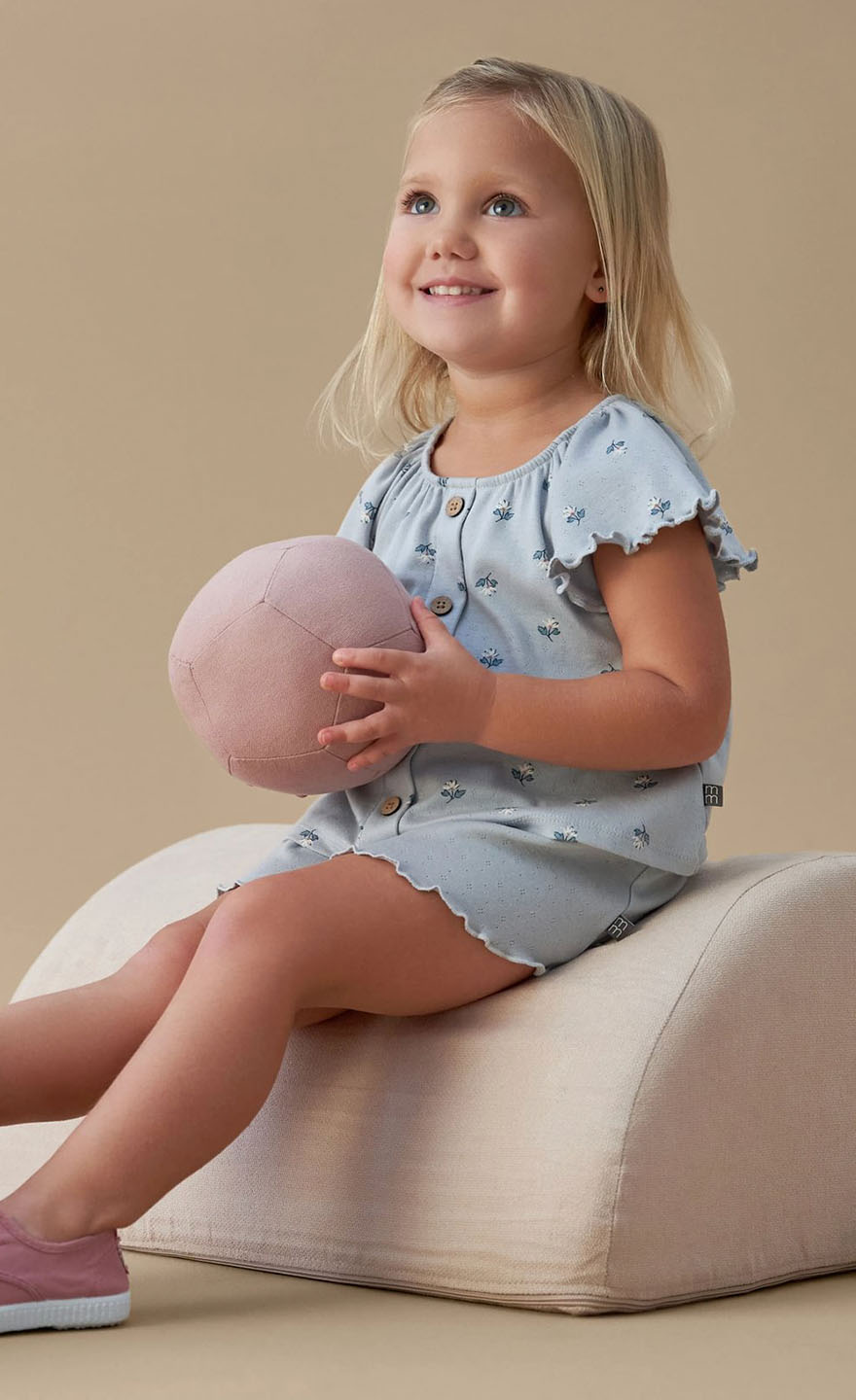 Young girl sitting on a beige cushion holding a pink ball against a beige background