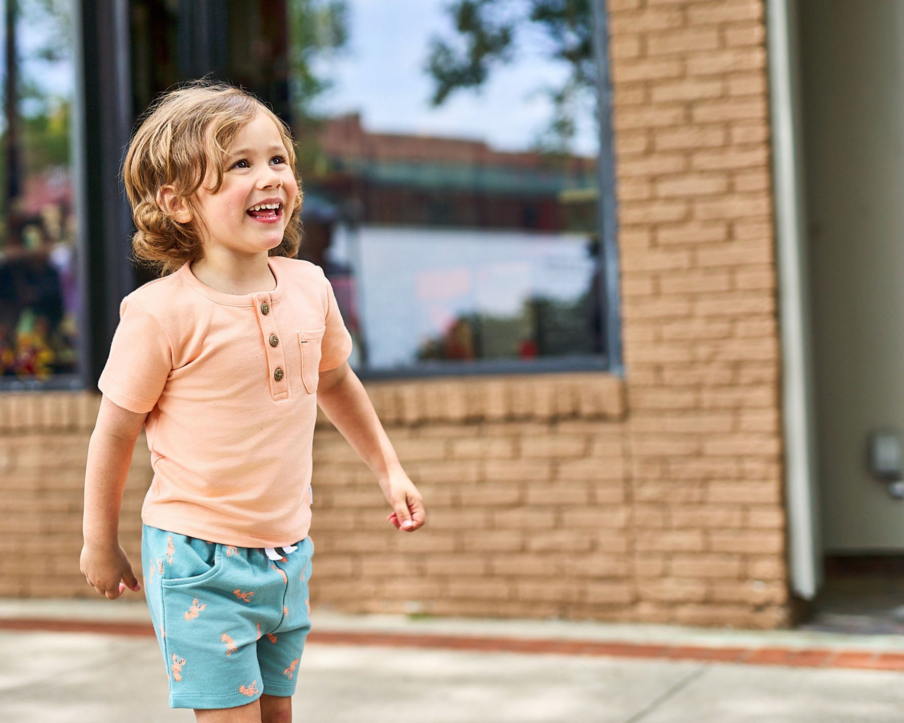 Child in a orange shirt and blue shorts standing outside a building