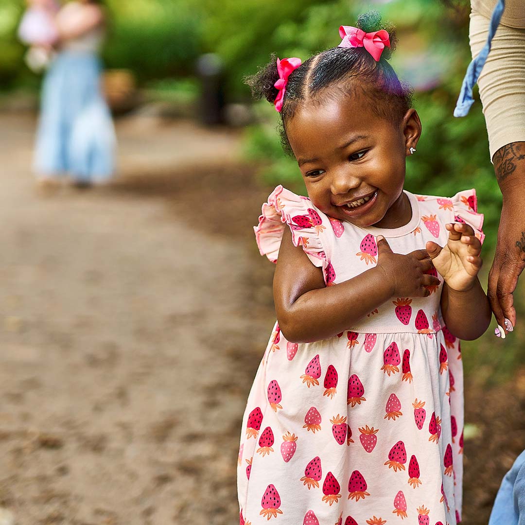 Child in a pink dress with strawberry pattern holding hands with an adult outdoors.