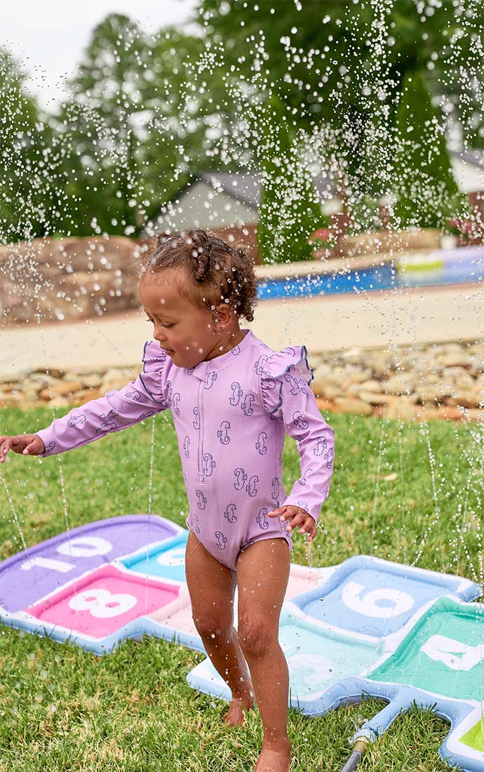 Child in a purple swimsuit playing with a sprinkler on a colorful hopscotch-style mat outdoors