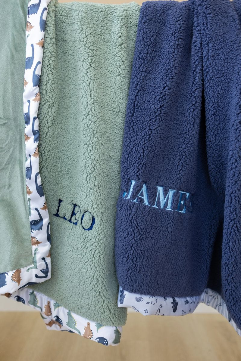 Two blankets, one green and one blue, with embroidered names 'Leo' and 'James' on a wooden surface.
