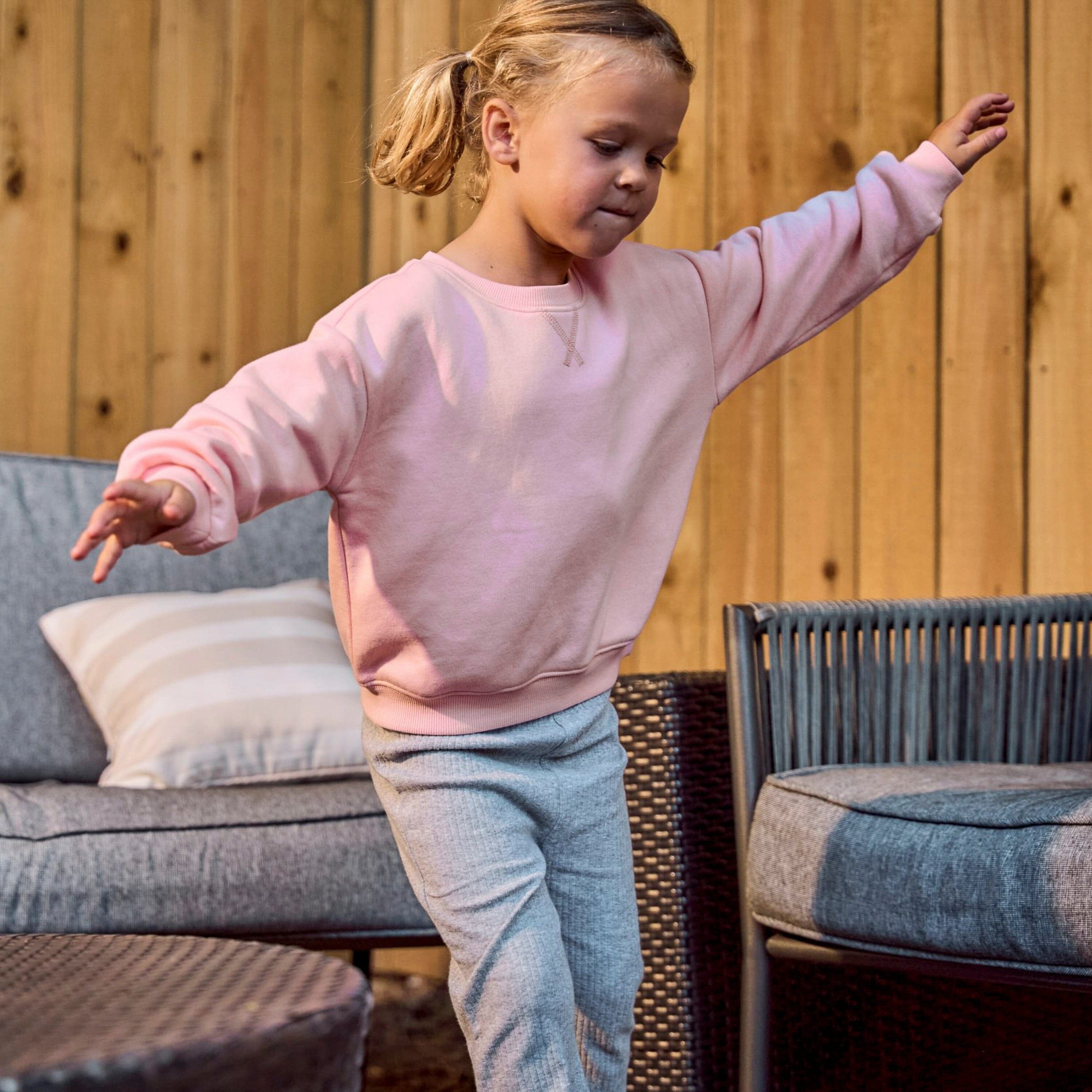 A young girl with blonde hair in a ponytail is wearing a light pink crewneck sweatshirt and light gray heathered sweatpants, posing with arms outstretched in front of a wooden wall and patio furniture.
