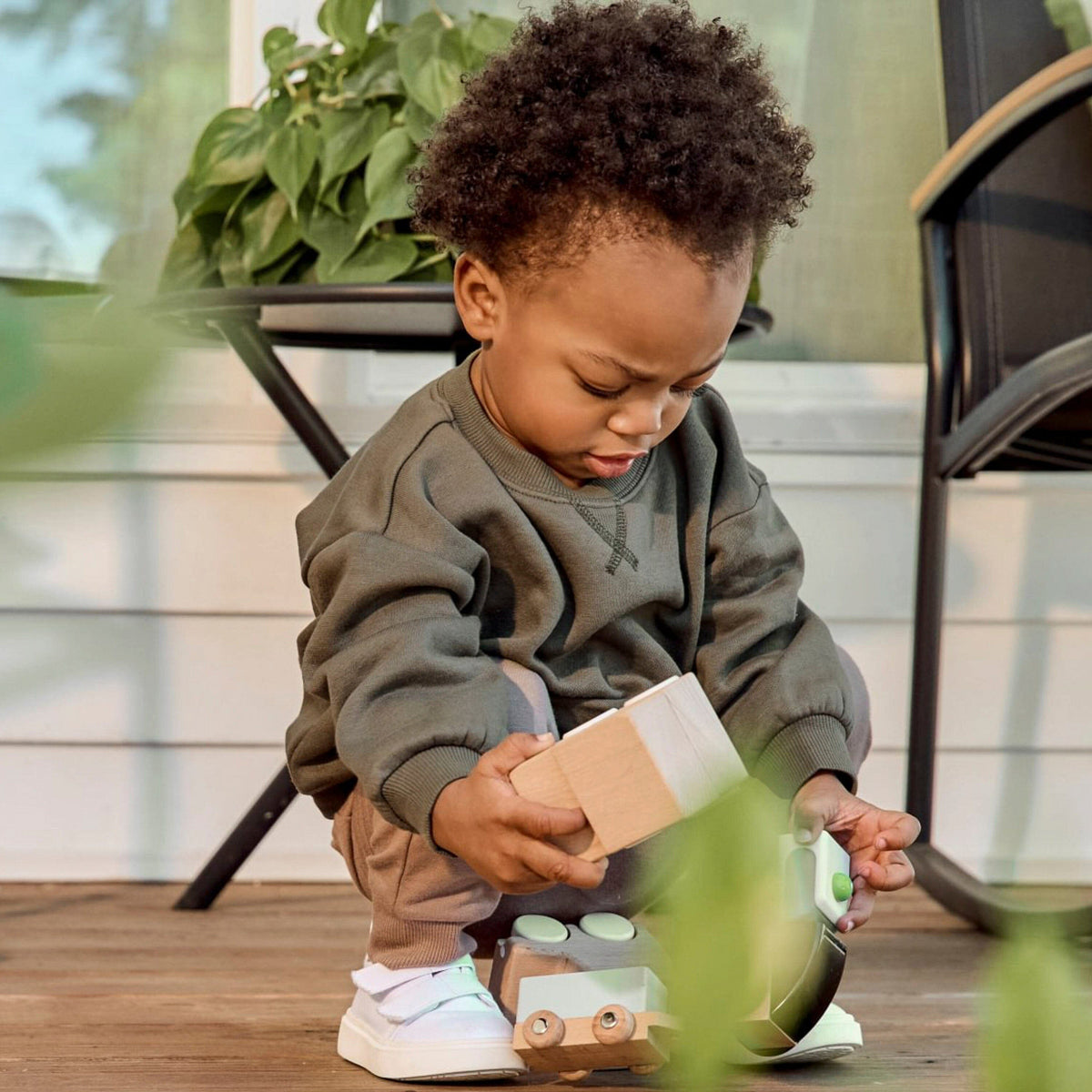 A toddler with curly dark hair sits on a wooden deck, wearing a dark olive green sweatshirt, tan pants, and white sneakers while playing with a wooden toy train.