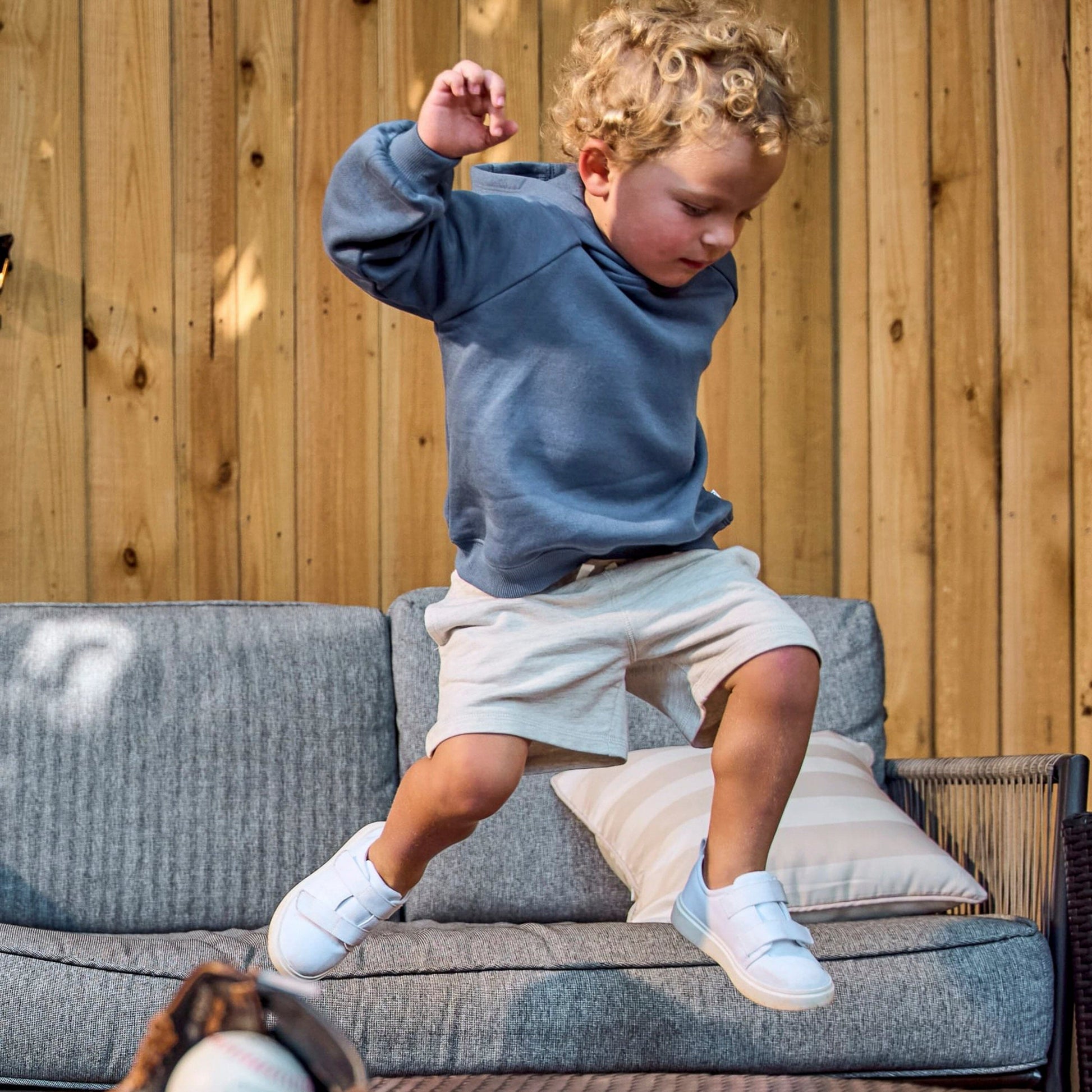 A young child with curly blonde hair wearing a slate blue hoodie, cream shorts, and white sneakers jumps onto a gray outdoor sofa in front of a wooden fence.
