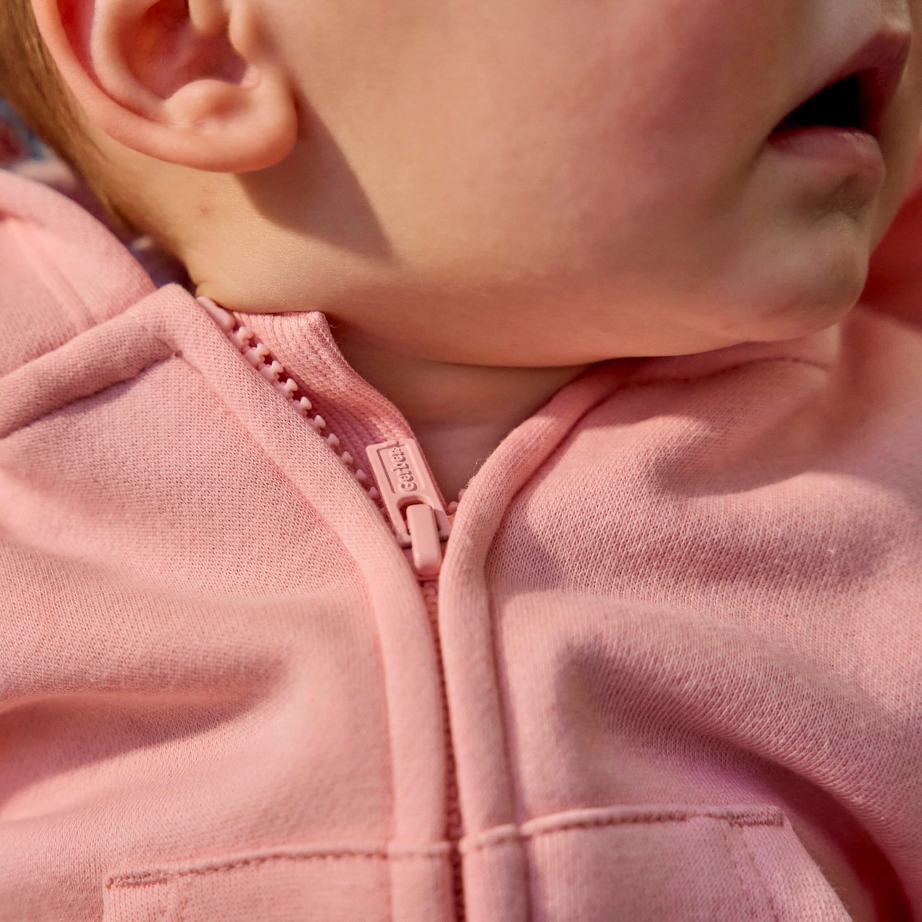 Close-up of a child wearing a dusty pink zip-up hoodie, showing the fabric texture, zipper pull, and part of the ear and mouth.