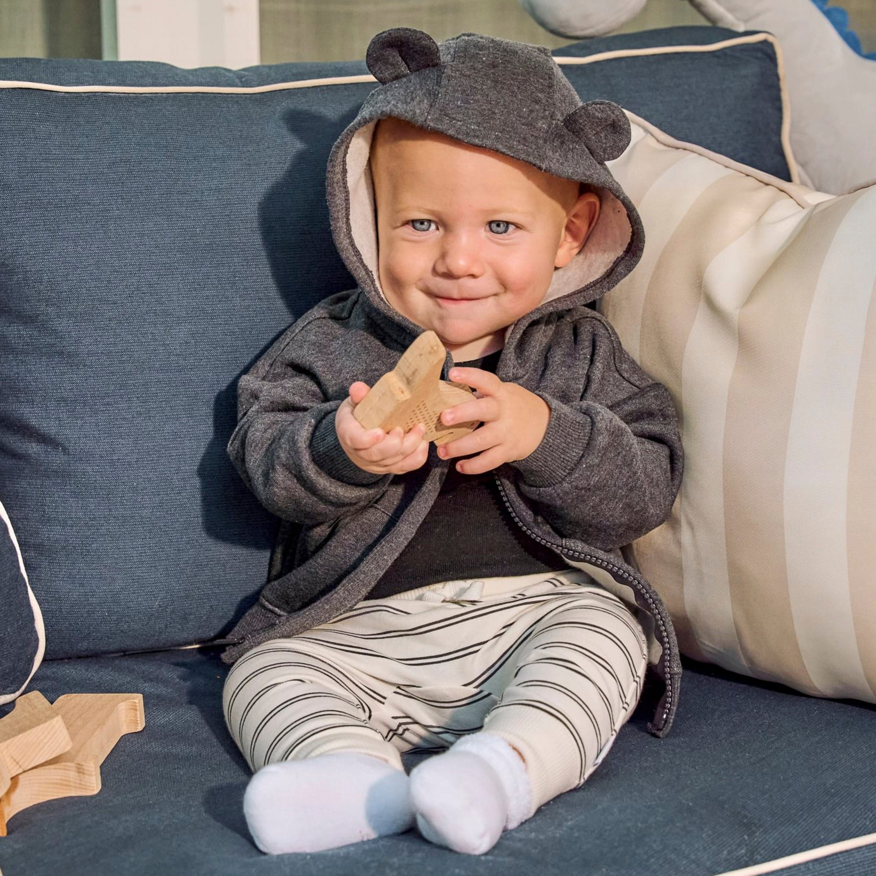 A baby sitting on a blue sofa wearing a dark gray hooded sweatshirt with bear ears and holding a wooden toy.