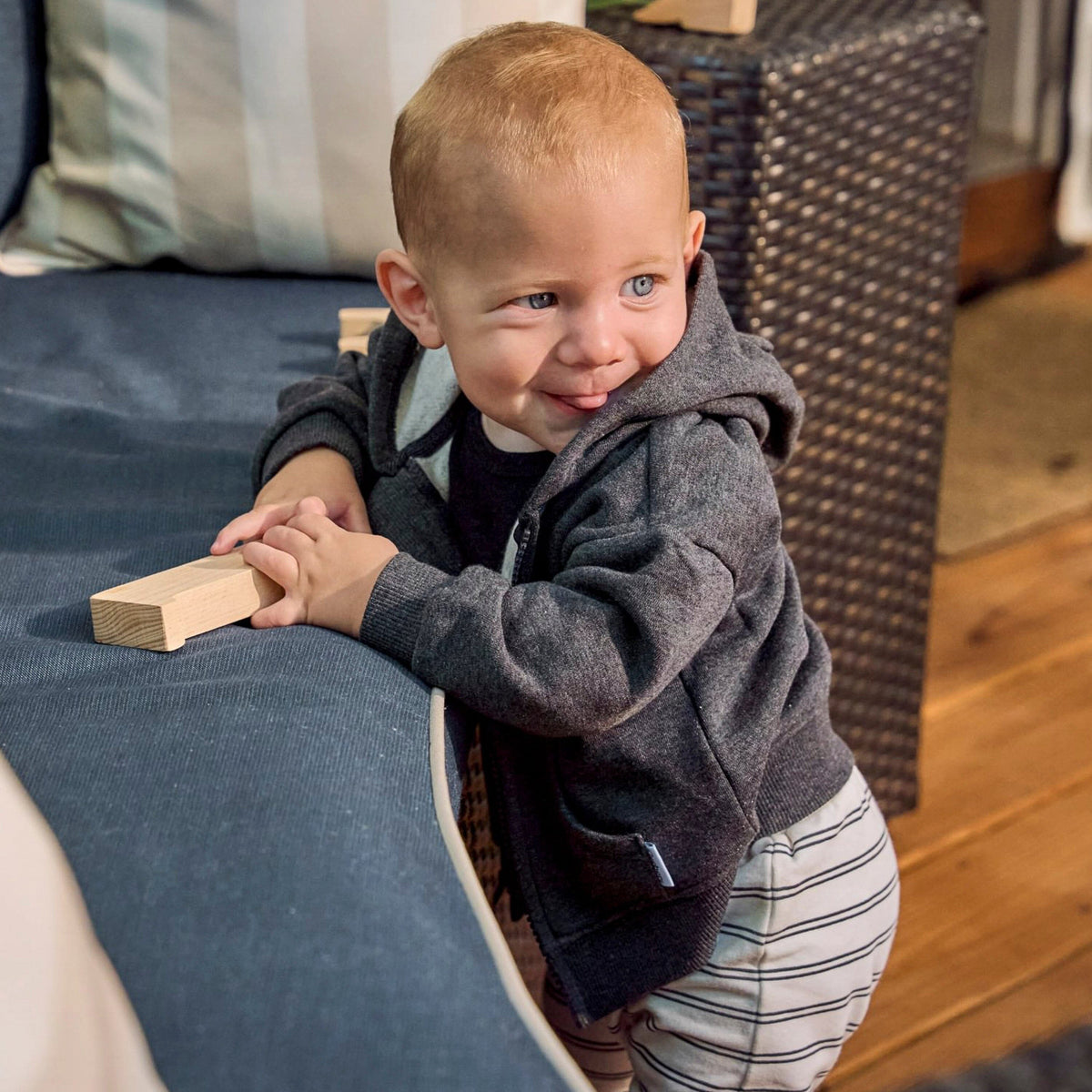 A smiling toddler with light hair stands on a blue sofa in a home setting, wearing a dark gray zip-up hoodie and striped pants, holding a small wooden block.