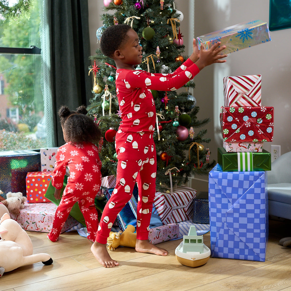 Two children in festive Christmas pajamas celebrate under a decorated tree. The standing child wears red pajamas with a white Santa Claus pattern and reaches for a wrapped gift, while the child on the left wears matching red pajamas with white snowflakes and looks at another gift.