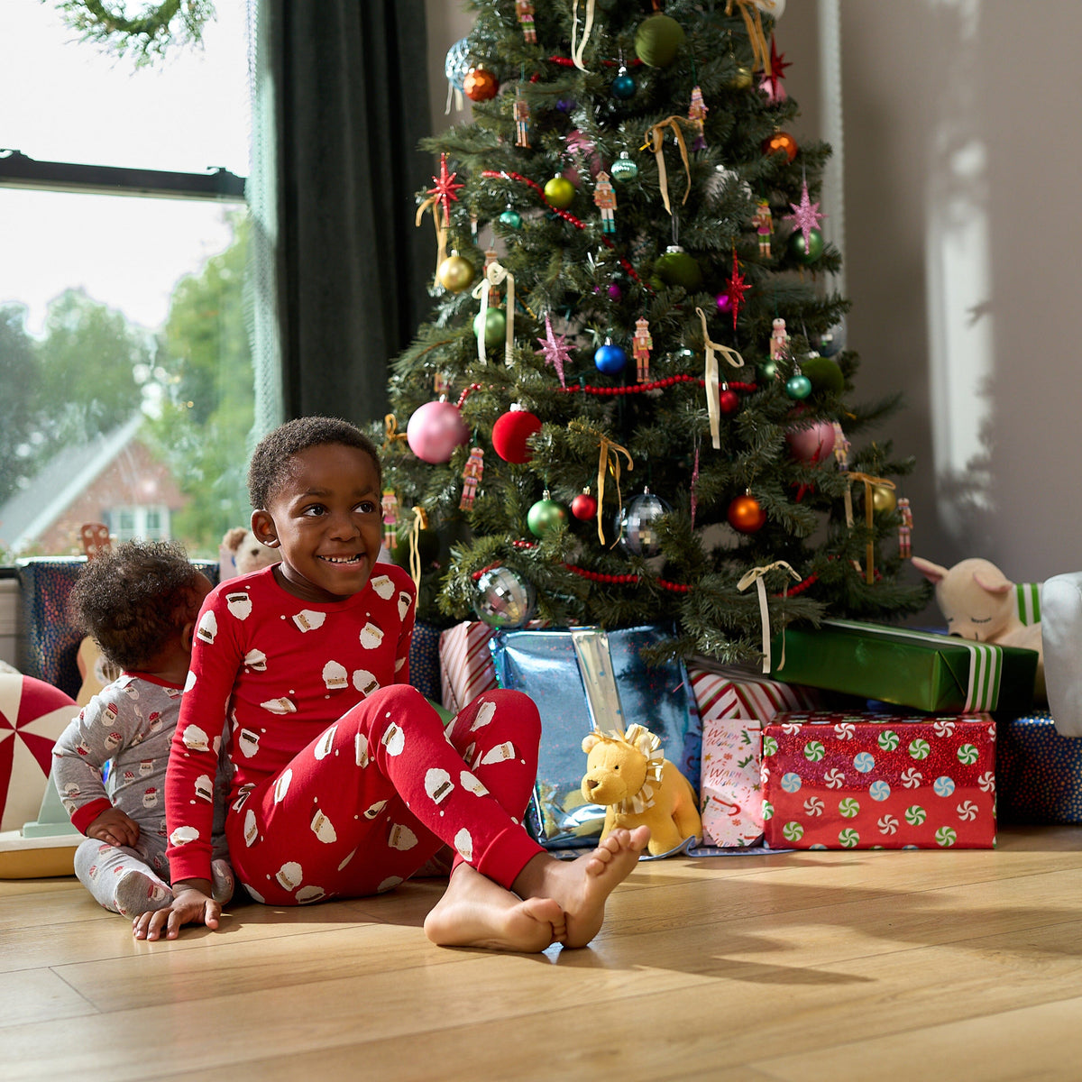 A young child wearing a red Christmas-themed pajama set with white patterns sits barefoot on a wooden floor in front of a decorated Christmas tree and wrapped presents.