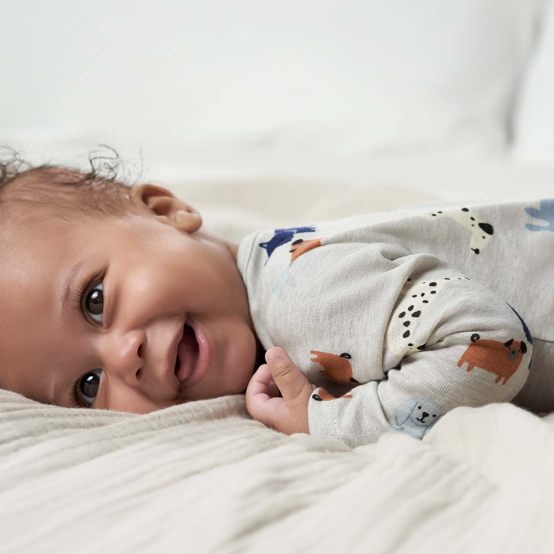 A happy baby lying on a soft cream-colored blanket, wearing a cozy animal-print onesie, smiling brightly at the camera. Showcasing the comfort and style we prioritize in every design.