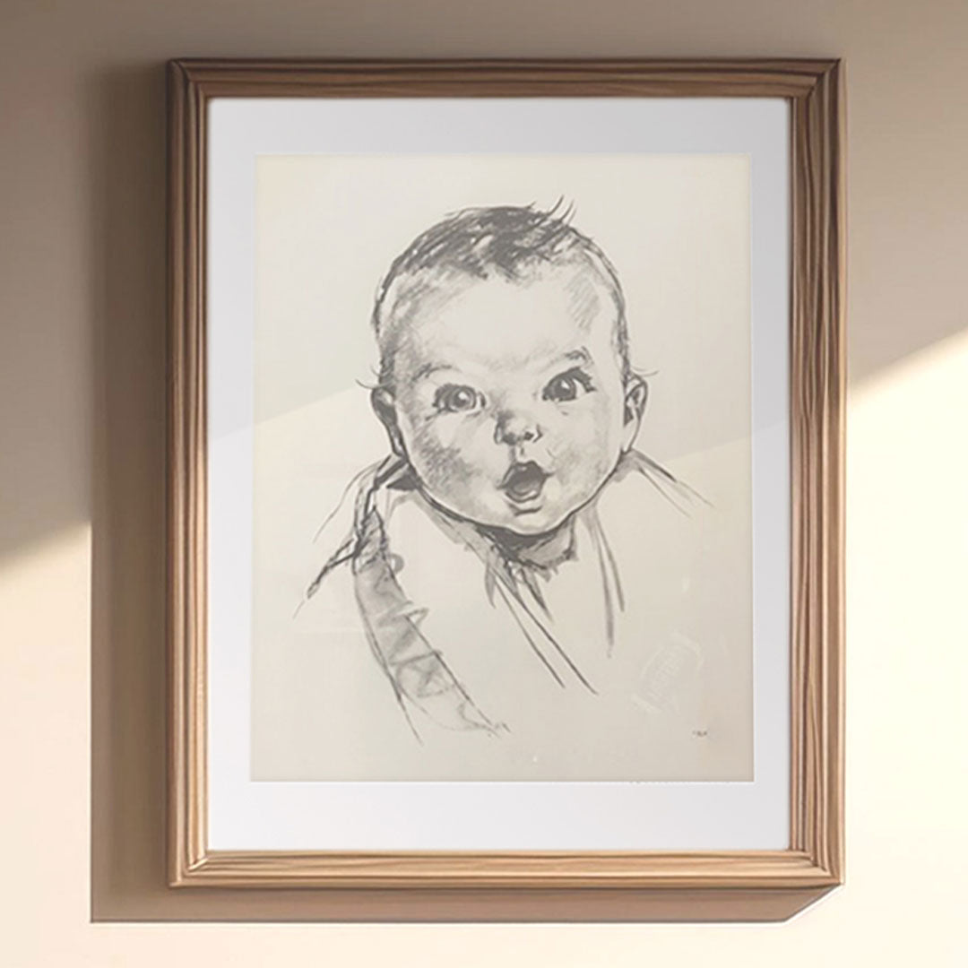 A framed black-and-white sketch of a baby with wide eyes and a playful expression, hanging on a softly lit wall. A nod to the timeless joy of childhood, featured on our ‘About Us’ page.