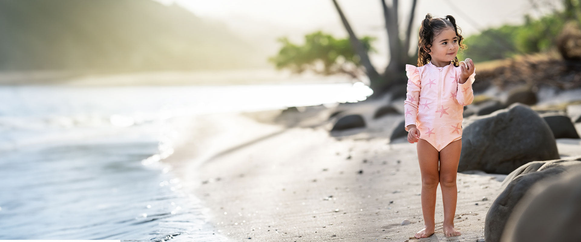 A young girl in a pink starfish print swimsuit stands thoughtfully on a sandy beach, with waves gently breaking in the background at sunset.
