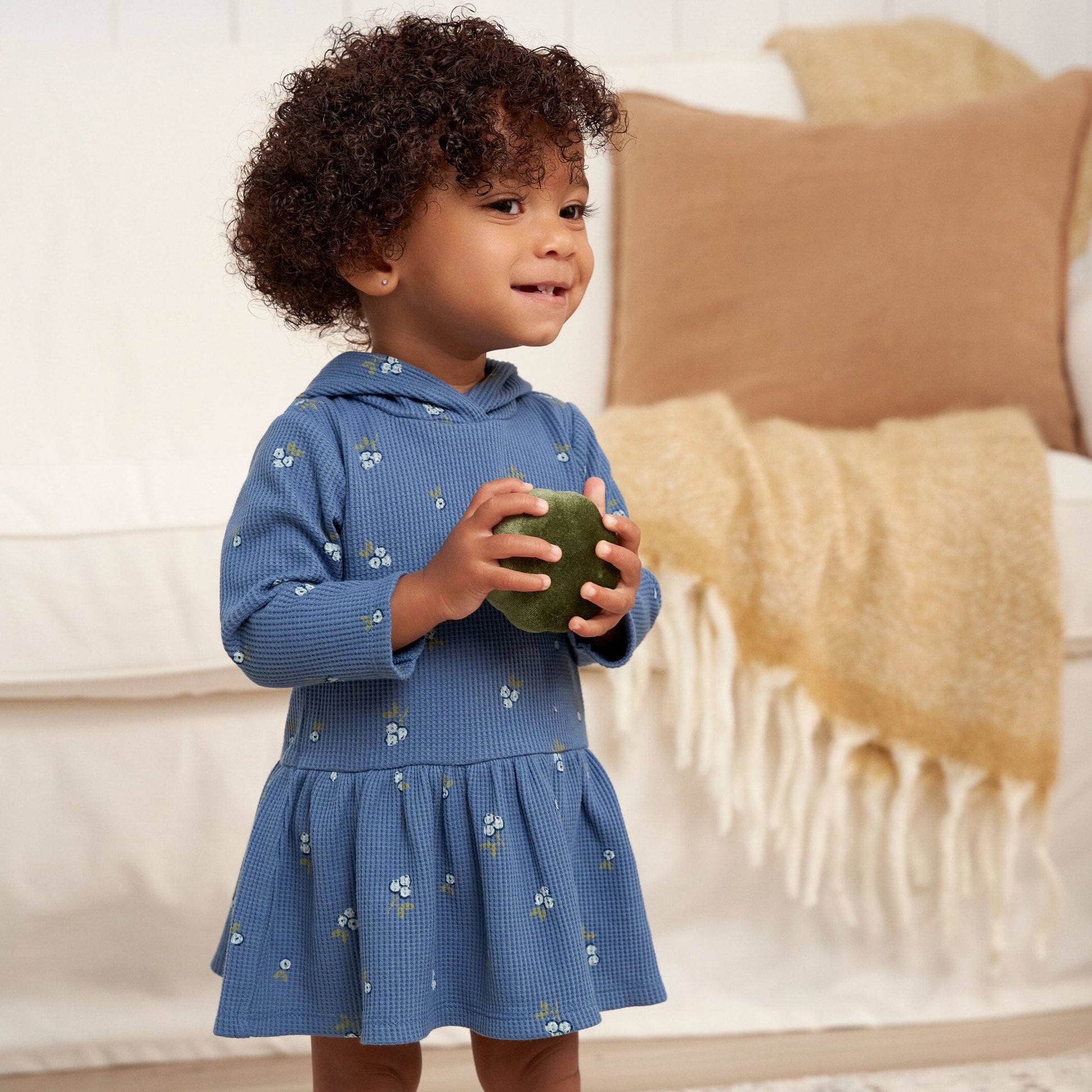 A young girl in a blue berry dress joyfully holds a bright red apple in her hands, smiling at the camera.