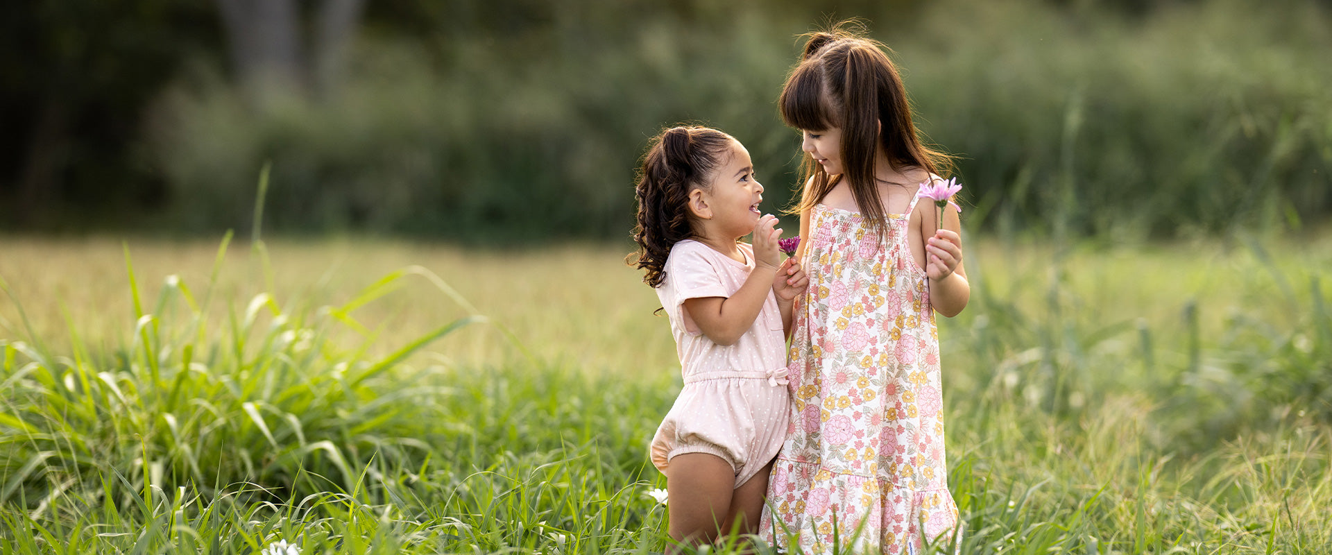 Two little girls wearing pink floral outfits are standing in a field together.