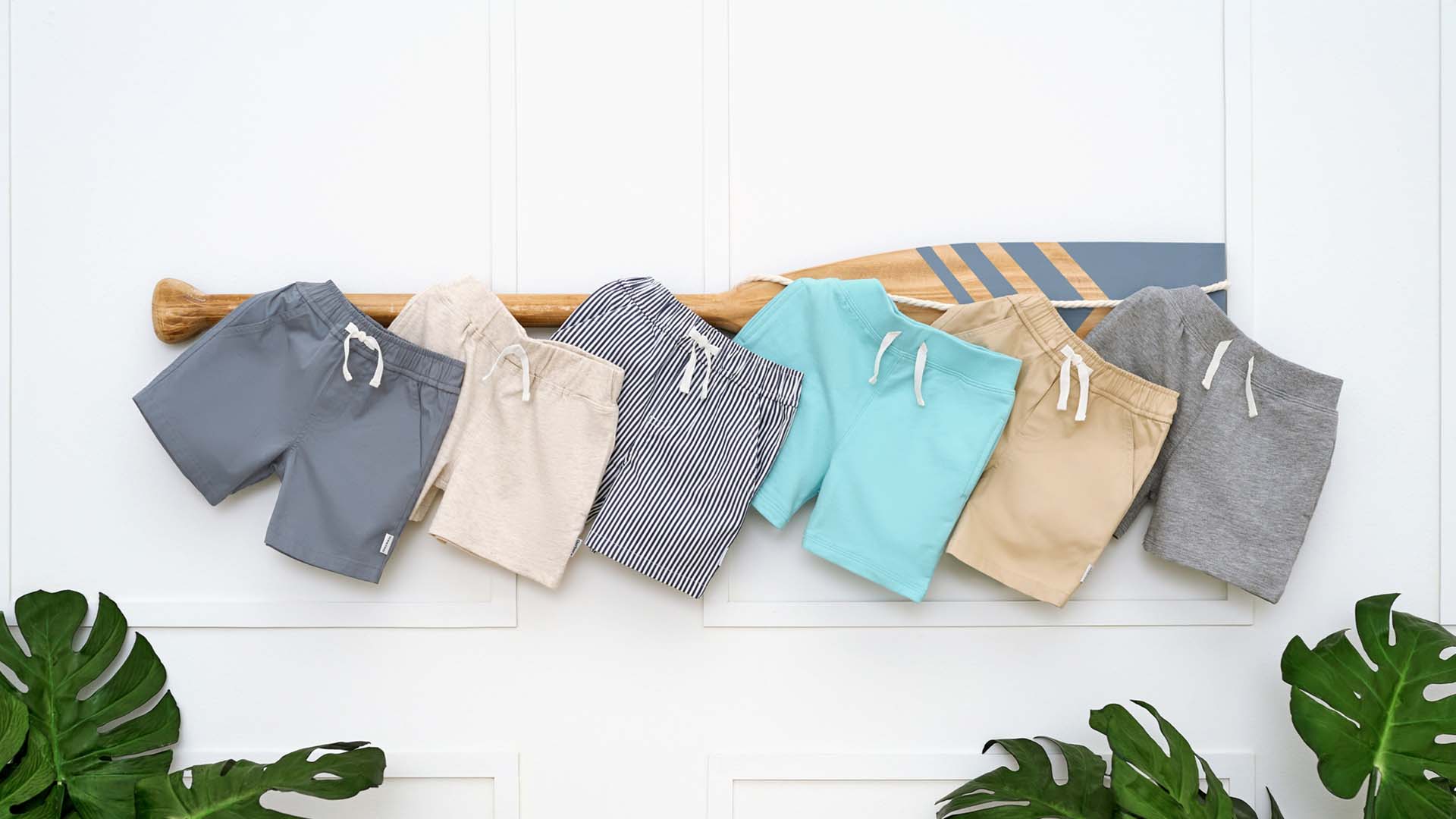 Variety of toddler boy's shorts in different colors and patterns, displayed in a row against a white backdrop with decorative plants on the sides.