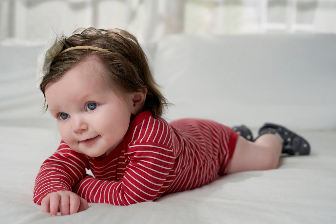 An infant dressed in holiday sleepwear, a red and white striped shirt, reclines on a bed.