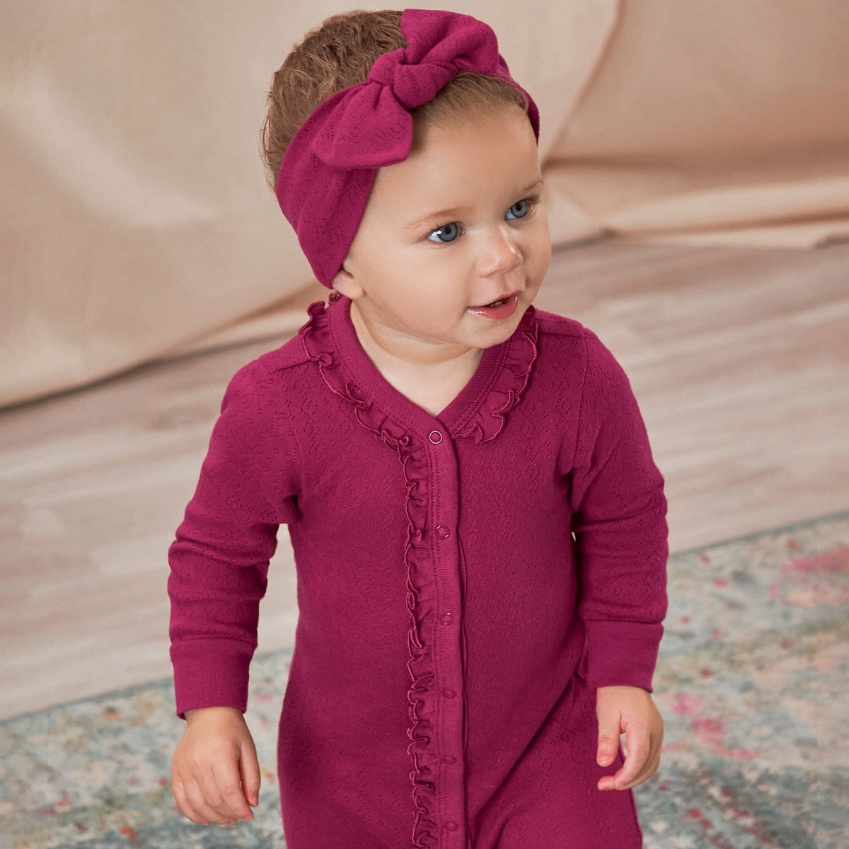 A baby girl wearing a pink onesie with a ruffle collar and a wine headband, looking to the right. She is standing on a rug with her hands at her side.  