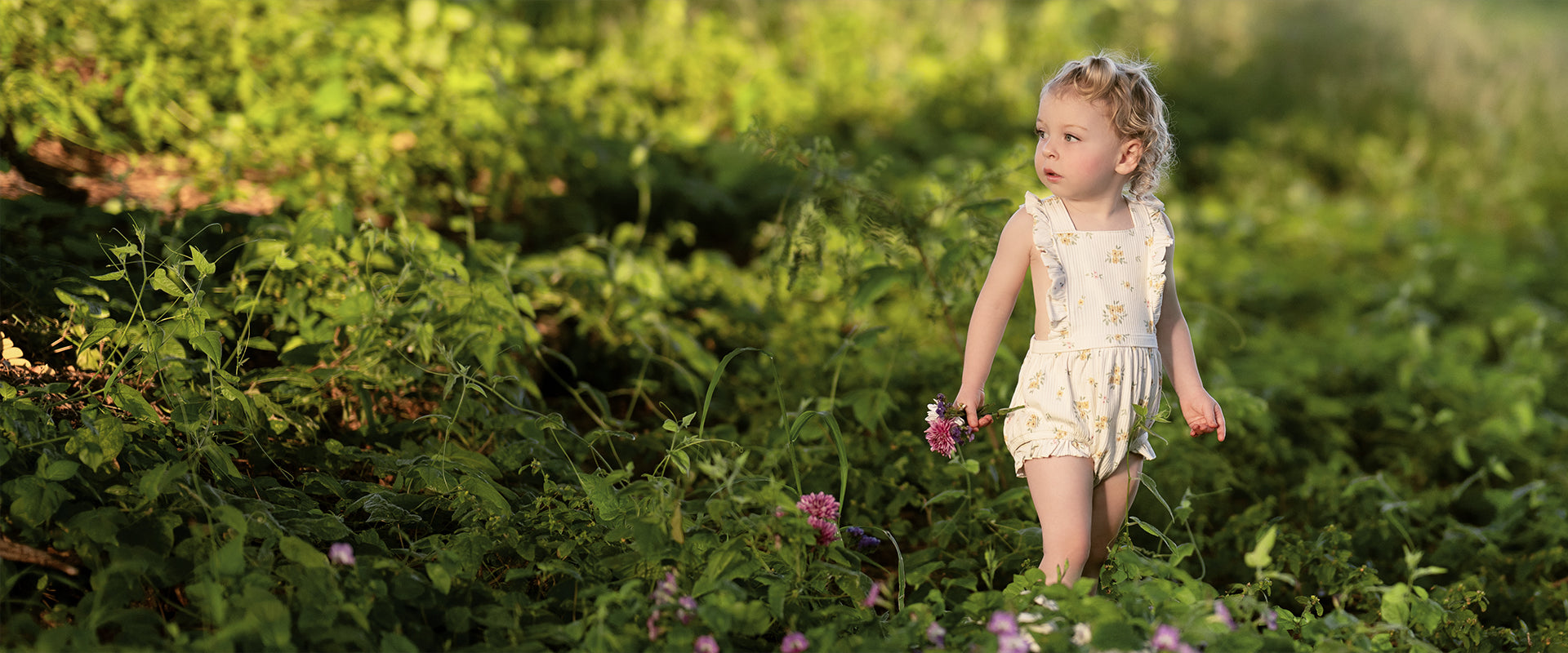 A little girl wearing a floral romper is standing in a field of flowers.